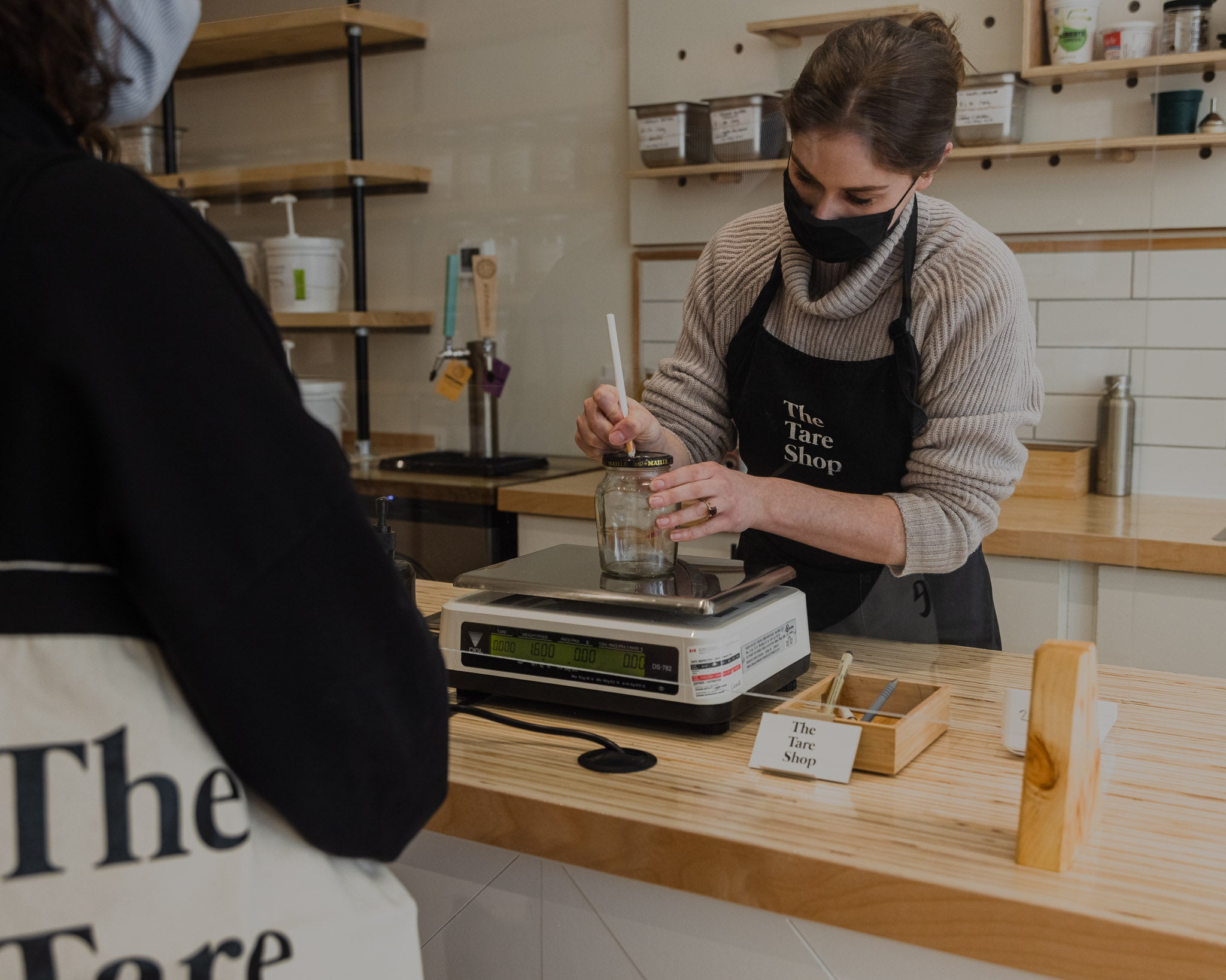 A staff member marks the lid of an empty jar with the jar's weight, while a customer waits behind plexiglass barrier.