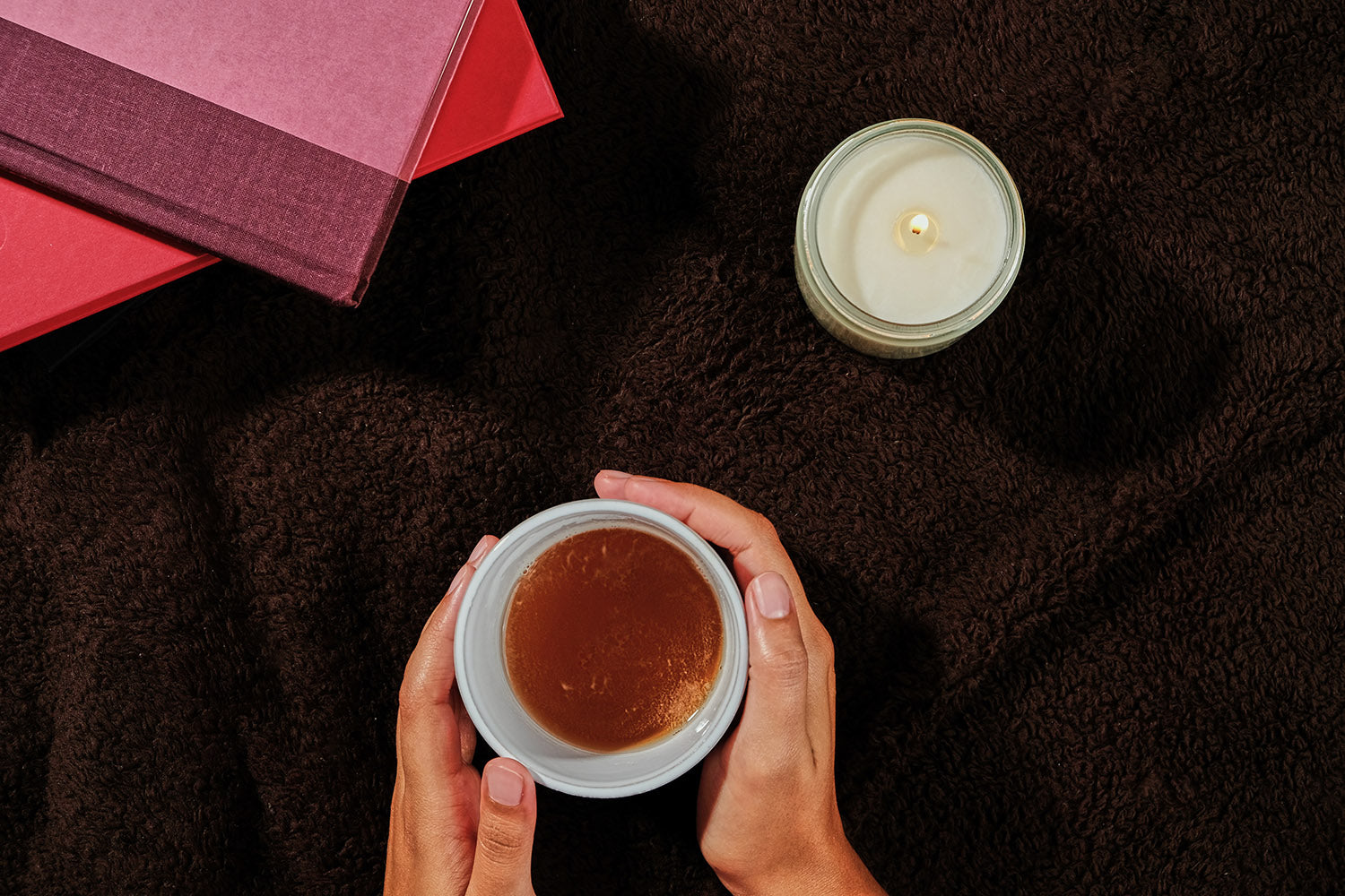 a hand holds a cup of tea with a candle and books in the background