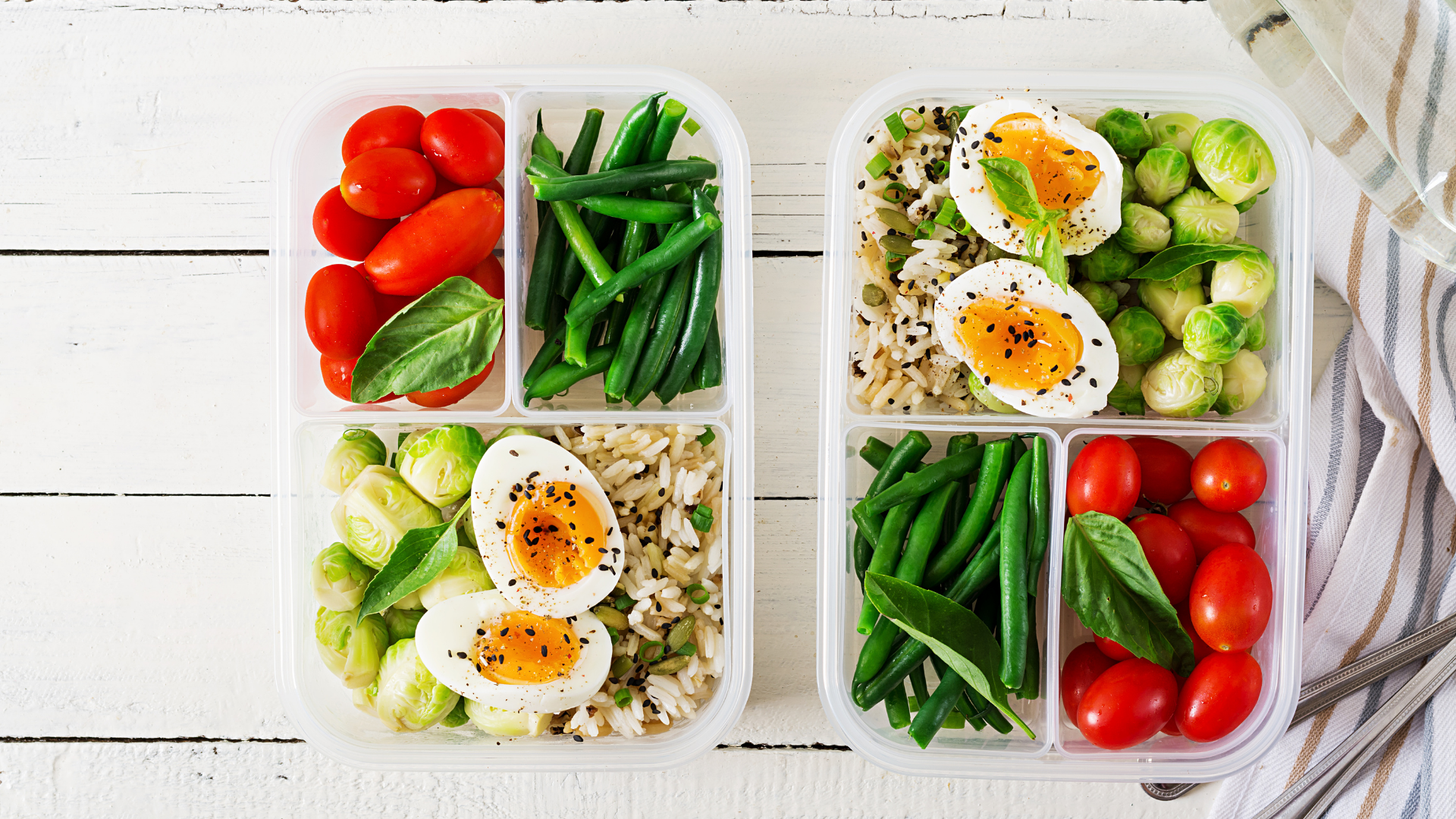 two containers with tomatoes, beans, hard boiled eggs, brussel sprouts and rice sit on a white wooden table.