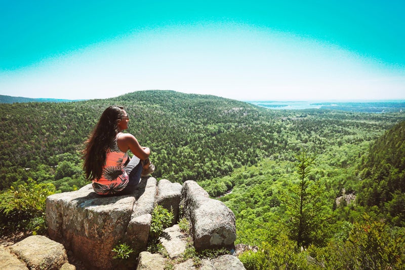a woman sits a the top of a hike on rocks starting out at the forest