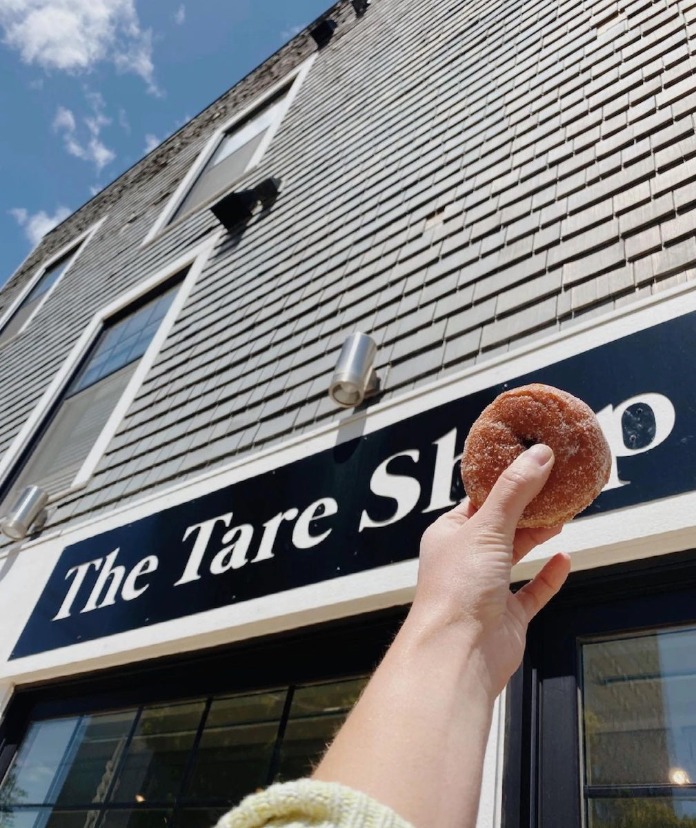 A hand holds up a donut over the "o" of "The Tare Shop" sign on a sunny day